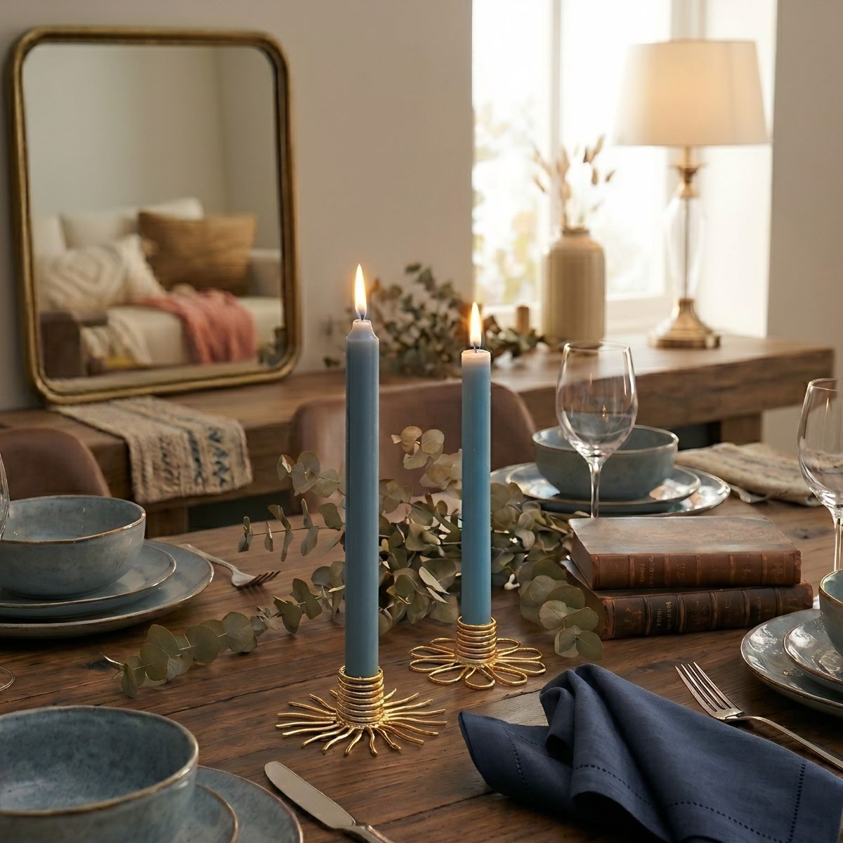 Blue candles in gold candle holders on a wooden dining table surface with a mirror in the background. Dining table is set with bowls, plates, napkins and wineglasses. 
