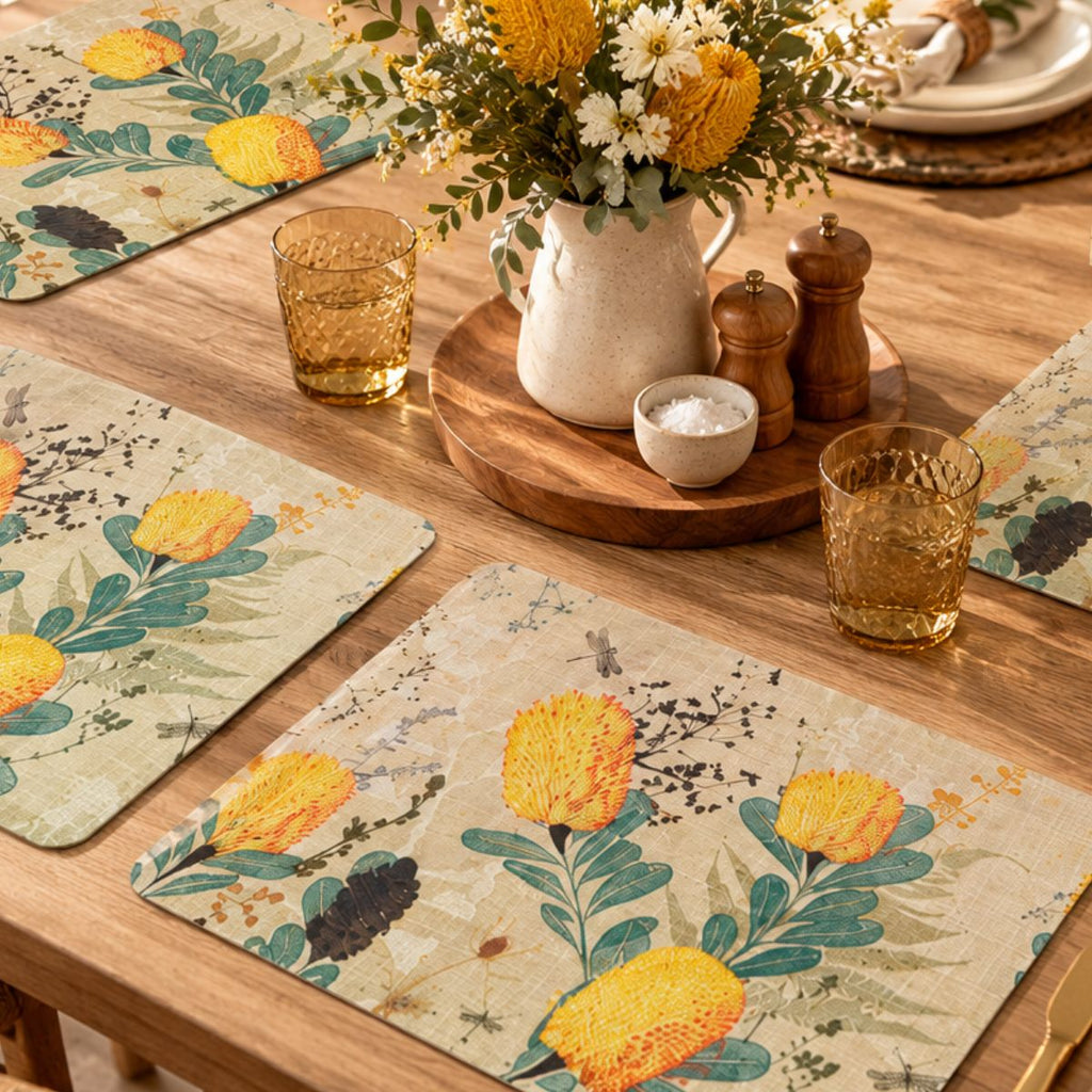 A styled dining table setting featuring rectangular placemats with a botanical floral print of yellow flowers and green leaves on a textured beige background. The table is also decorated with amber glass tumblers, a ceramic pitcher of white and yellow flowers, wooden salt and pepper mills, and a round wooden serving tray.