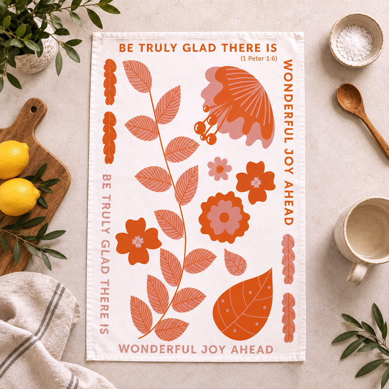 A white kitchen tea towel with orange floral and botanical illustrations and the text 'BE TRULY GLAD THERE IS (1 Peter 1:4) WONDERFUL JOY AHEAD' is laid flat on a light beige surface, surrounded by lifestyle props including lemons, a cutting board, a mug, and small bowls.