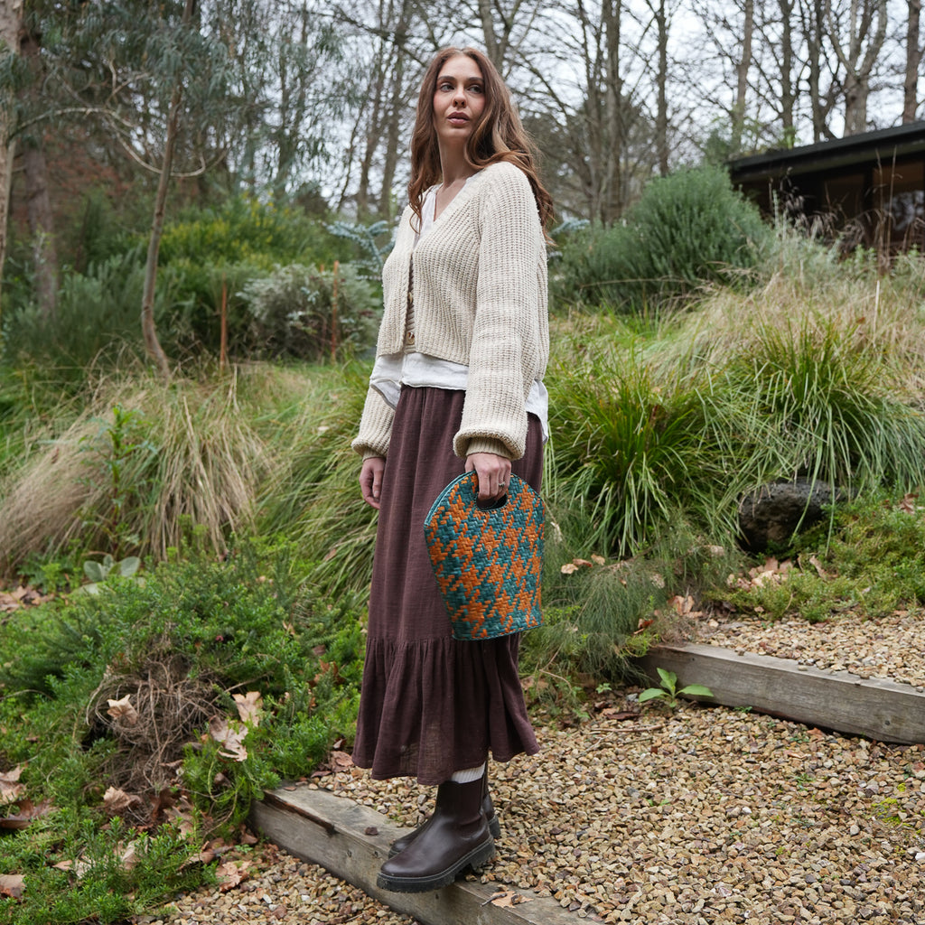 A woman stands outdoors in a garden setting wearing a cream ribbed cardigan, a dark brown tiered midi skirt, and dark brown boots, holding a blue and orange patterned handbag.