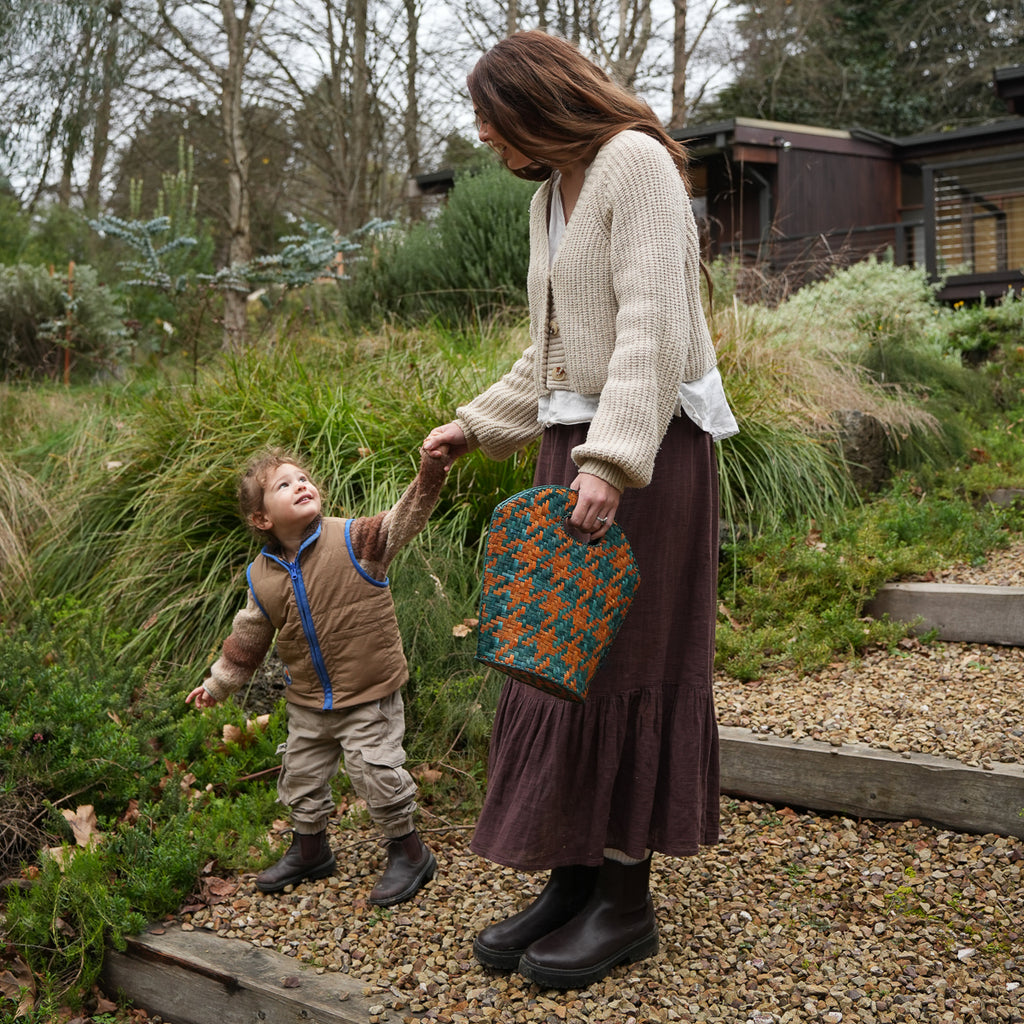 A woman and a young boy walking outdoors on a gravel path, holding hands. The woman is wearing a cream knit cardigan, a dark brown skirt, and black boots, while holding a teal and orange patterned handbag.