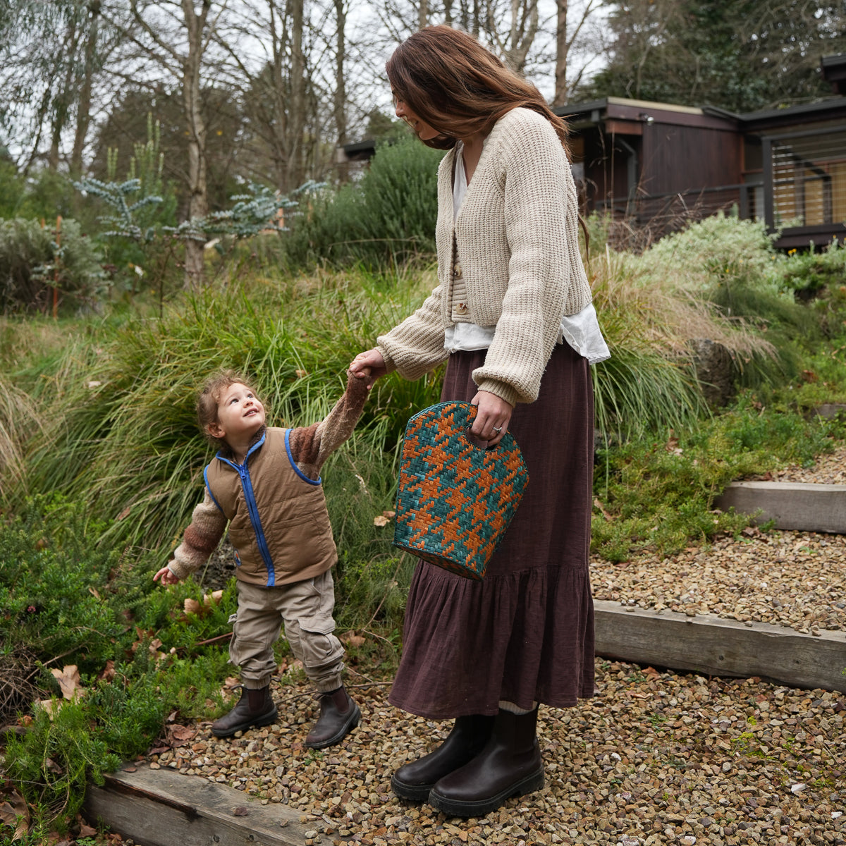 A woman and a young boy walking outdoors on a gravel path, holding hands. The woman is wearing a cream knit cardigan, a dark brown skirt, and black boots, while holding a teal and orange patterned handbag.