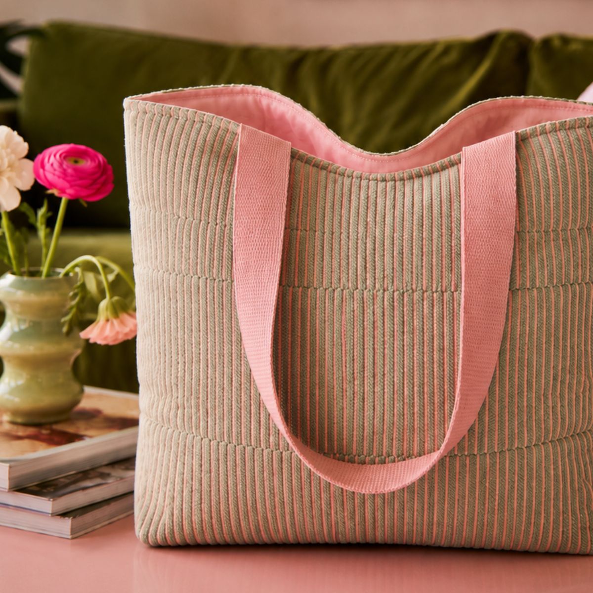 Beige tote bag with pink handles on a pink surface with a green cushion and flowers in the background.