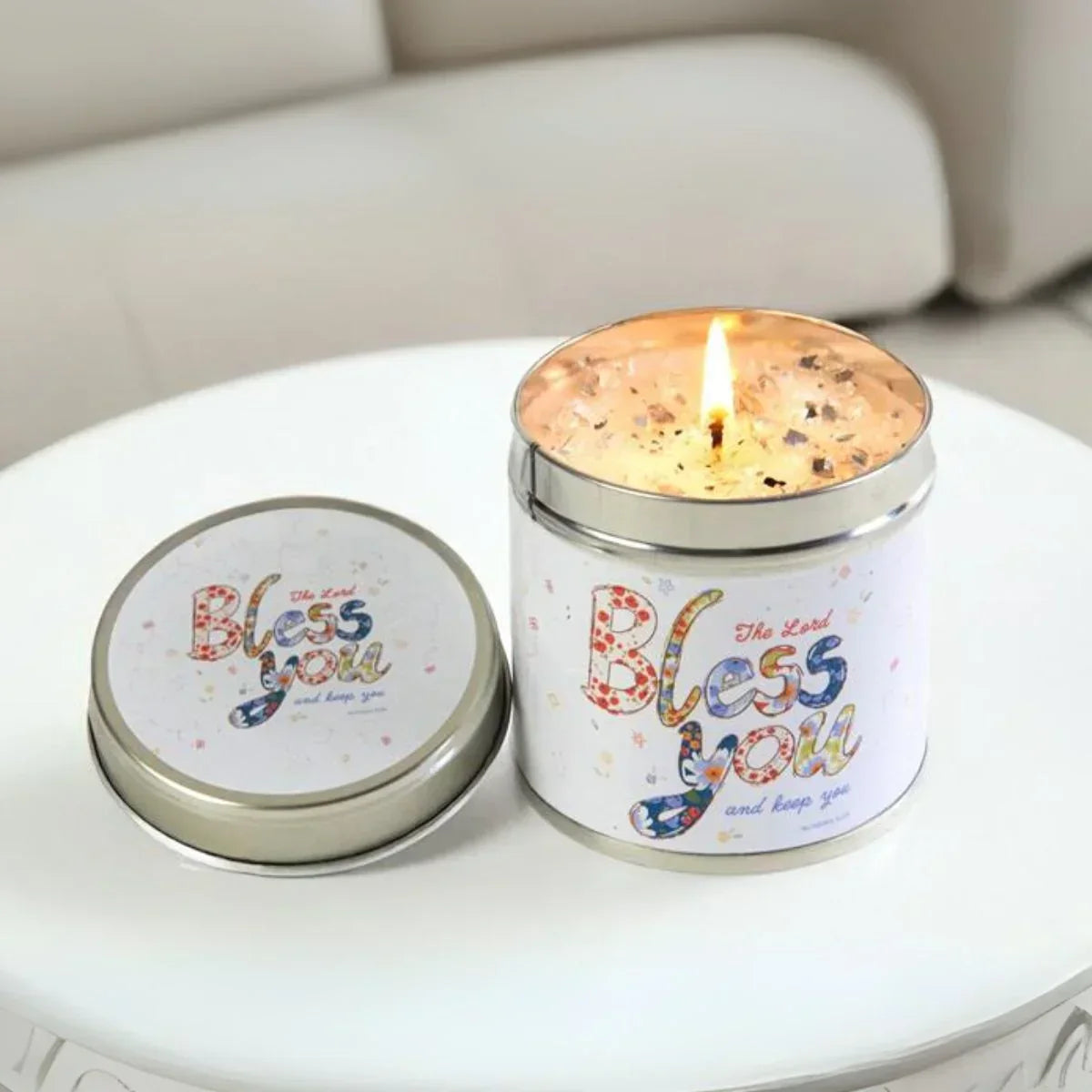 Lit candle in a decorative tin with 'Bless you' text on a white surface.