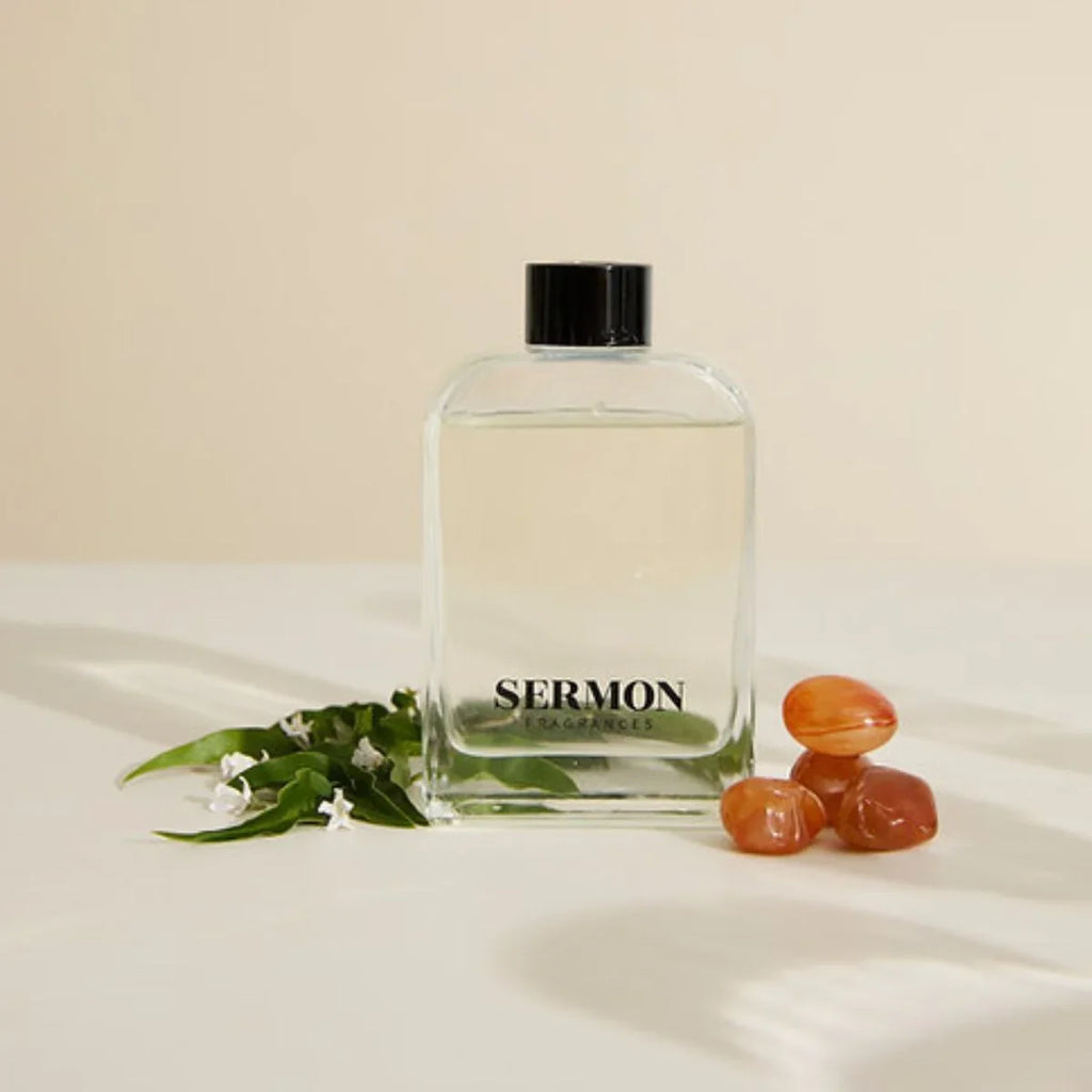 Clear bottle labeled 'SERMON' with black cap on a beige surface with green leaves and orange stones.