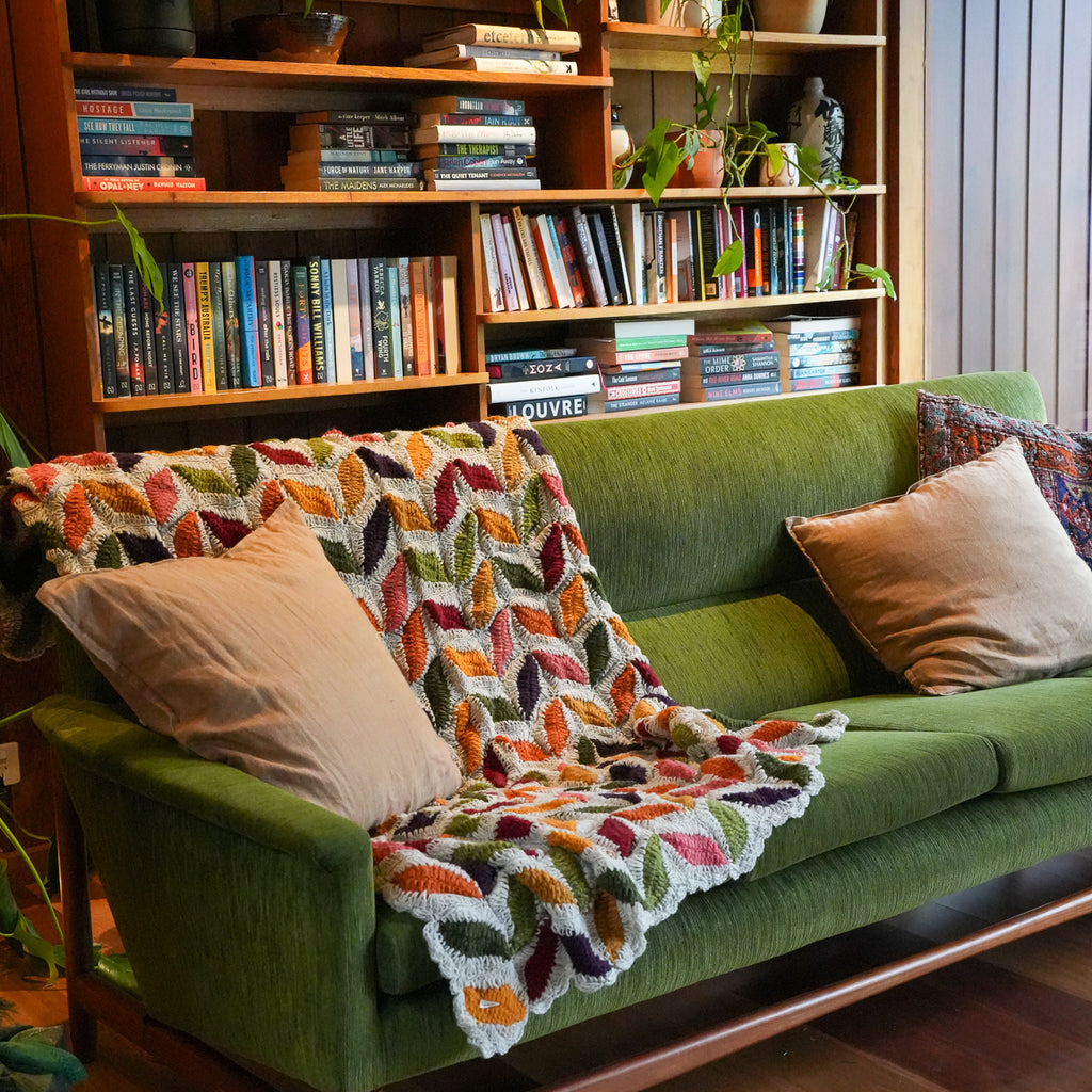 A cozy green sofa in a living room setting, styled with a colorful crocheted throw blanket featuring a leaf pattern and neutral beige pillows. A large wooden bookshelf filled with books stands behind the sofa.