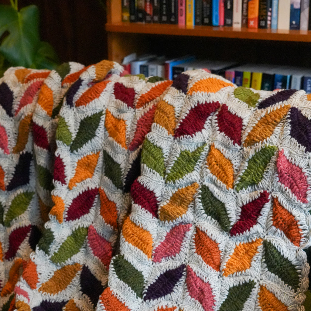 A close-up of a textured, multicolored woven throw blanket draped over a chair, featuring a repeating leaf pattern in orange, green, red, pink, and purple on a cream background, with a blurred bookshelf in the background.