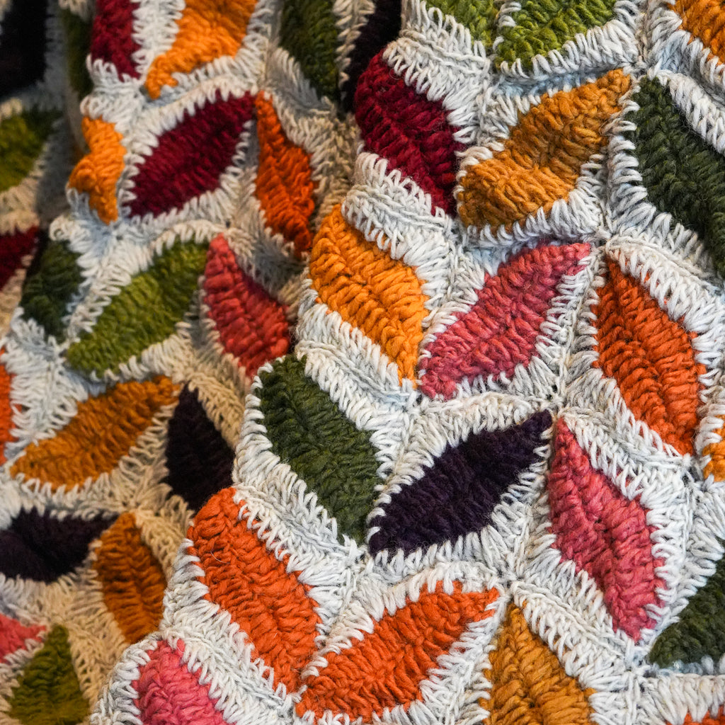 A close-up of a textured, chunky crocheted blanket featuring a geometric leaf pattern in multiple colors including orange, pink, green, red, and dark purple on a white base.