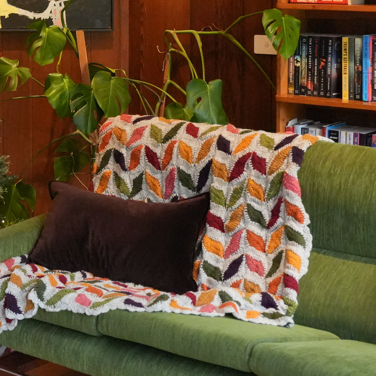 A multicolor chevron-patterned knit throw blanket draped over a green sofa, styled with a dark brown pillow in a cozy living room setting with a wooden bookshelf and houseplants in the background.