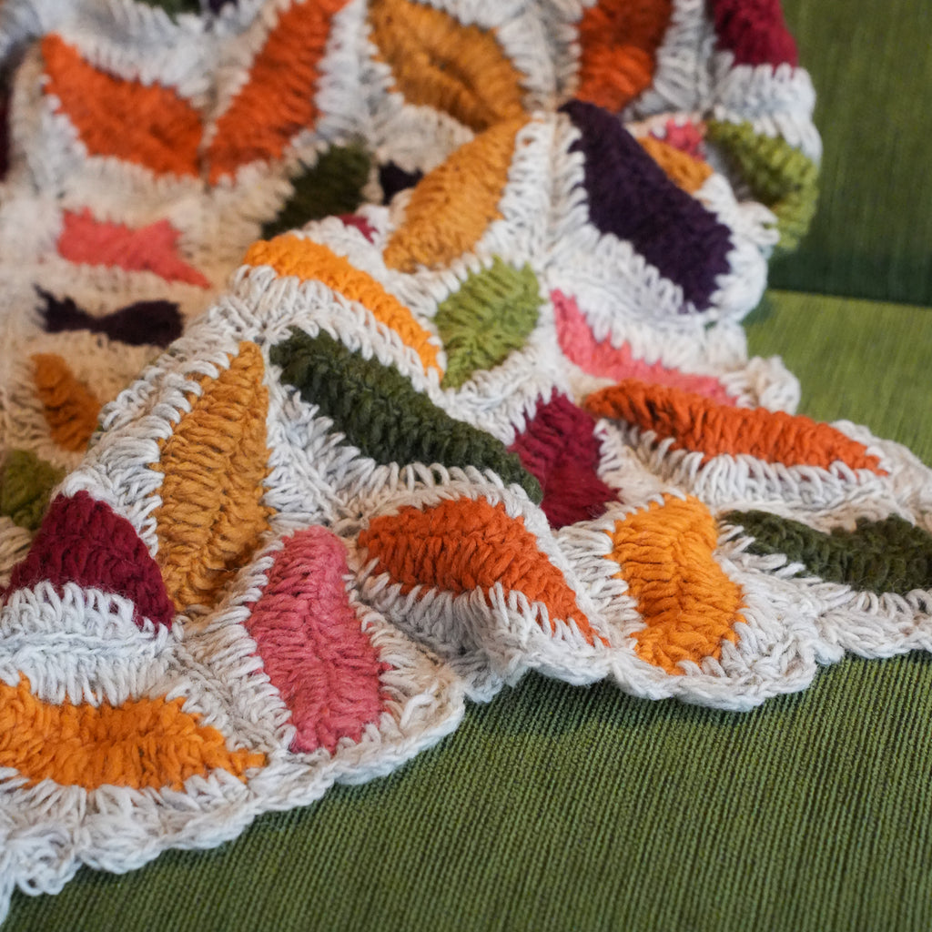 A close-up of a chunky crocheted throw blanket with textured, raised leaf-like shapes in orange, pink, purple, green, and mustard yellow, draped over a green textured chair.