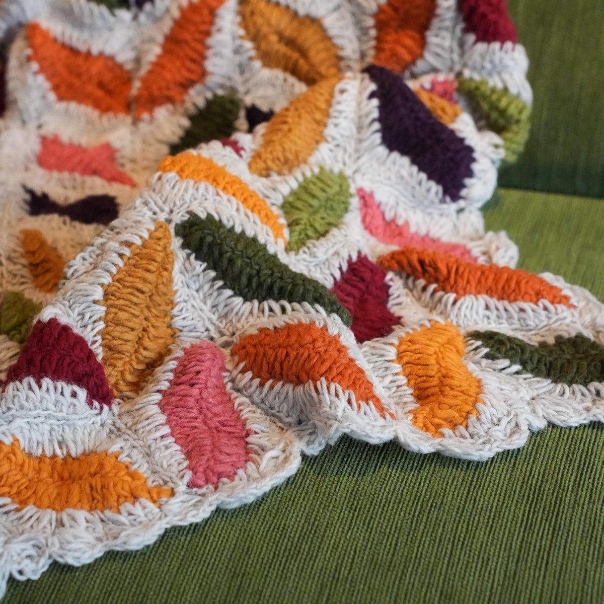 A close-up of a chunky crocheted throw blanket with textured, raised leaf-like shapes in orange, pink, purple, green, and mustard yellow, draped over a green textured chair.