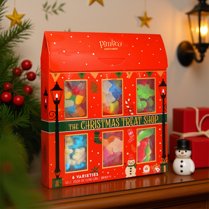 A red house-shaped Pimlico Christmas Gummy Treat Box sitting on a wooden mantelpiece, surrounded by festive decorations including pine branches with red berries, a lit wall sconce, and a small snowman figurine.