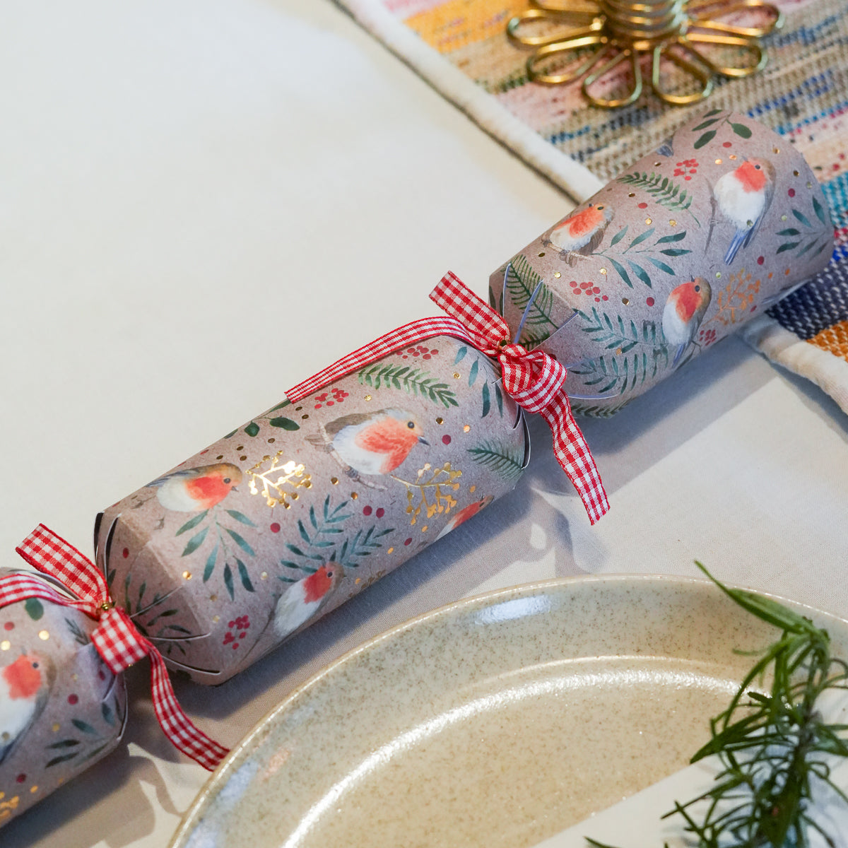 A Christmas cracker ornament wrapped in gray wrapping paper with a robin bird and leaf pattern, tied with red and white gingham ribbons, shown on a tabletop with a ceramic plate and decorative gold flower.