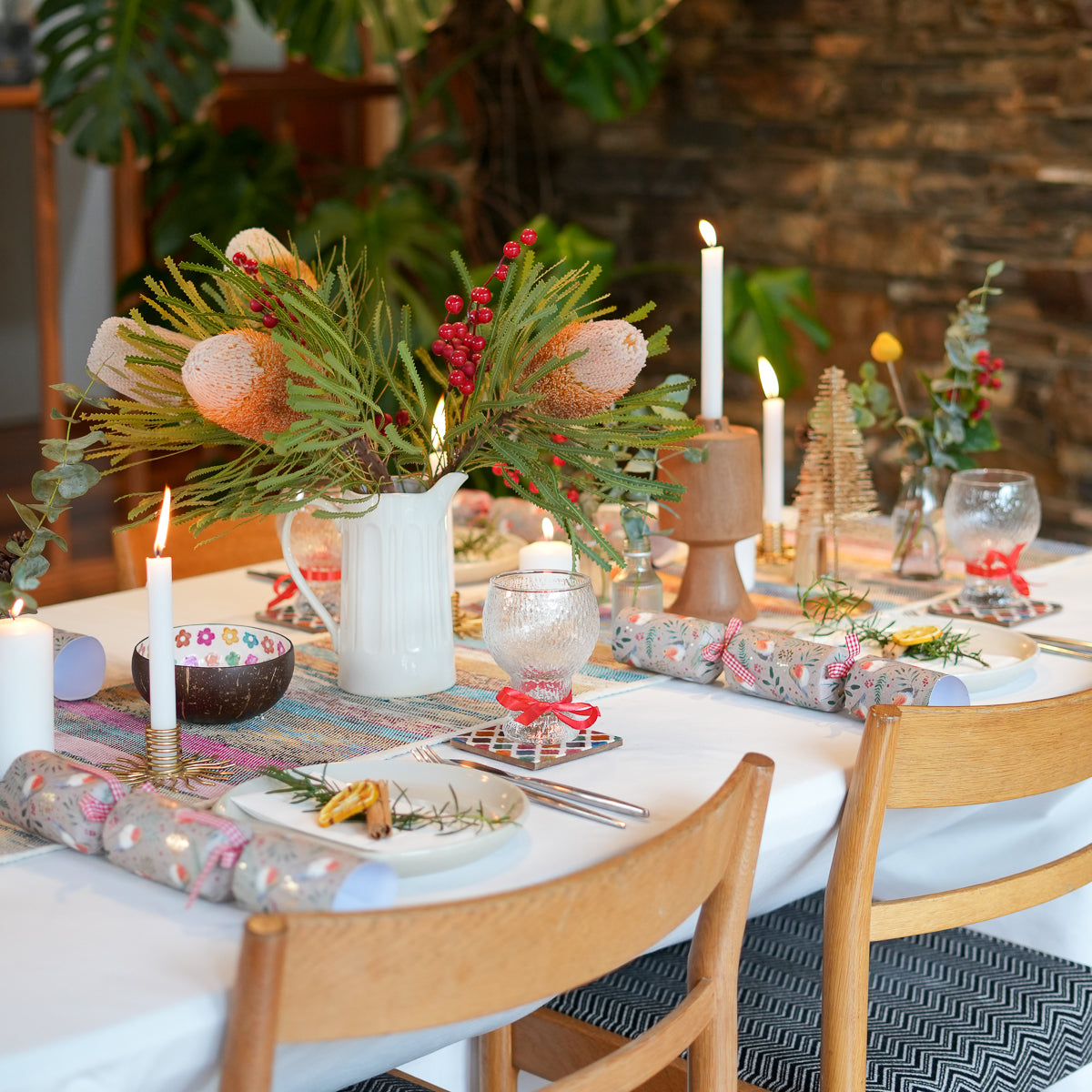 A festive dining table decorated for the holidays, featuring a white pitcher vase filled with green pine branches, pink protea flowers, and red berries, alongside lit white taper candles, patterned tableware, and wrapped gift rolls on a white tablecloth.