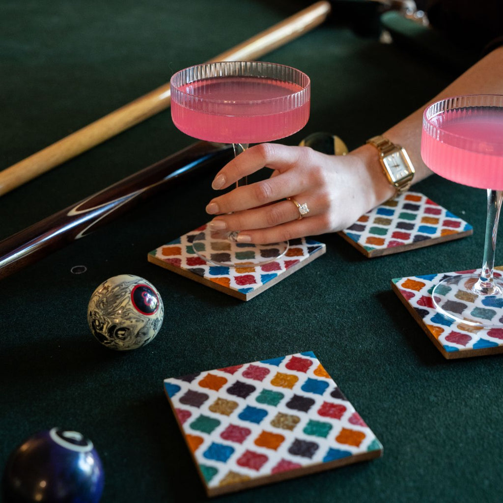 Hand holding a pink cocktail glass on a pool table with pool cues and balls.