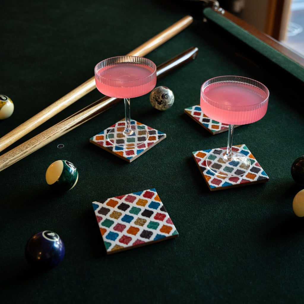 Two pink cocktails on colorful square coasters with pool balls and a cue on a green felt background.