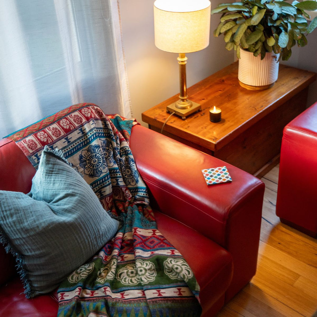 Red couch with a colorful blanket and blue pillow, wooden side table with lamp and candle, plant in the background.