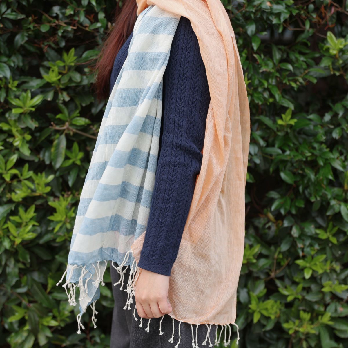 Person wearing a blue and white striped scarf and a peach-colored scarf against a green leafy background