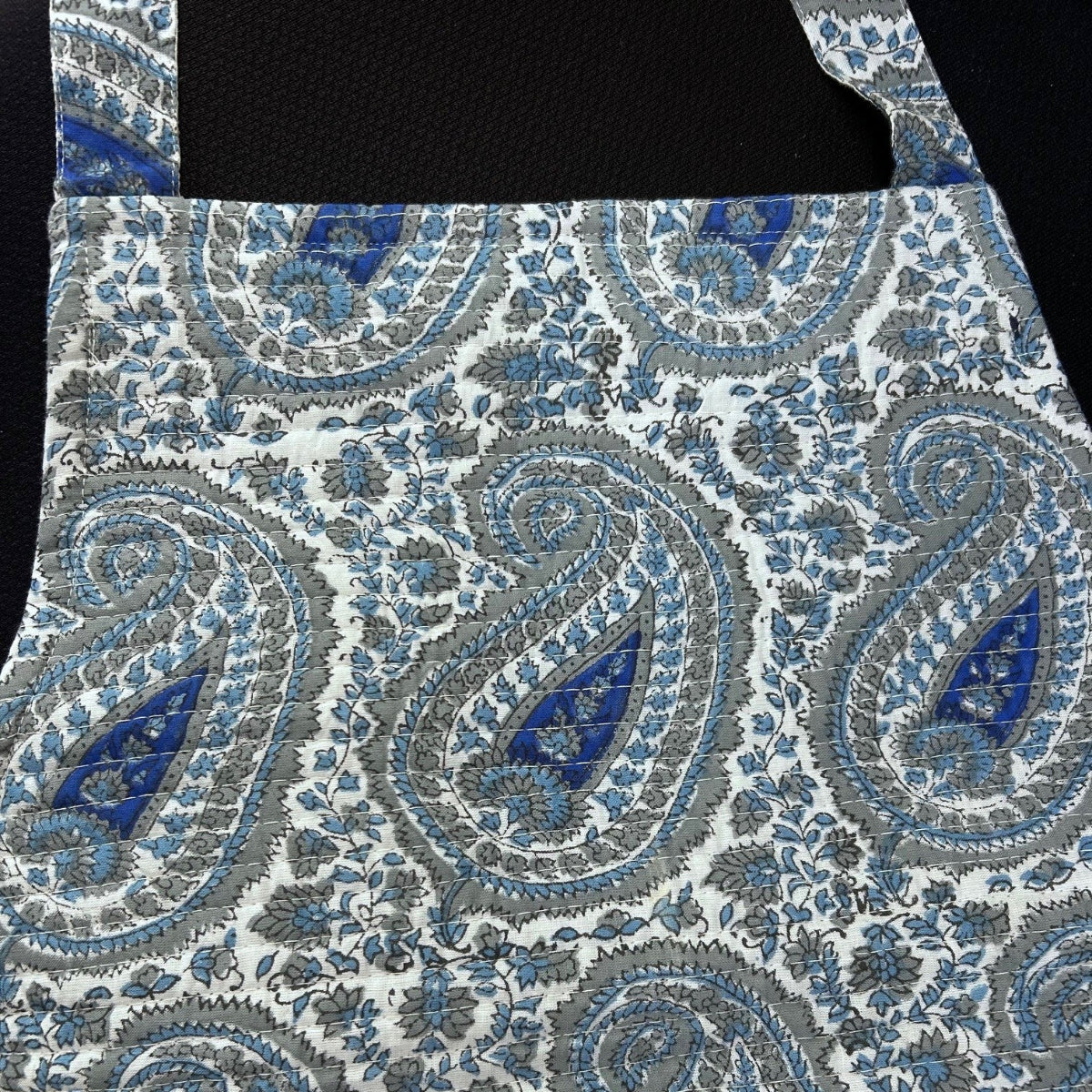 A close-up of a fabric product, likely an apron, featuring a detailed blue, gray, and white paisley pattern against a black background.