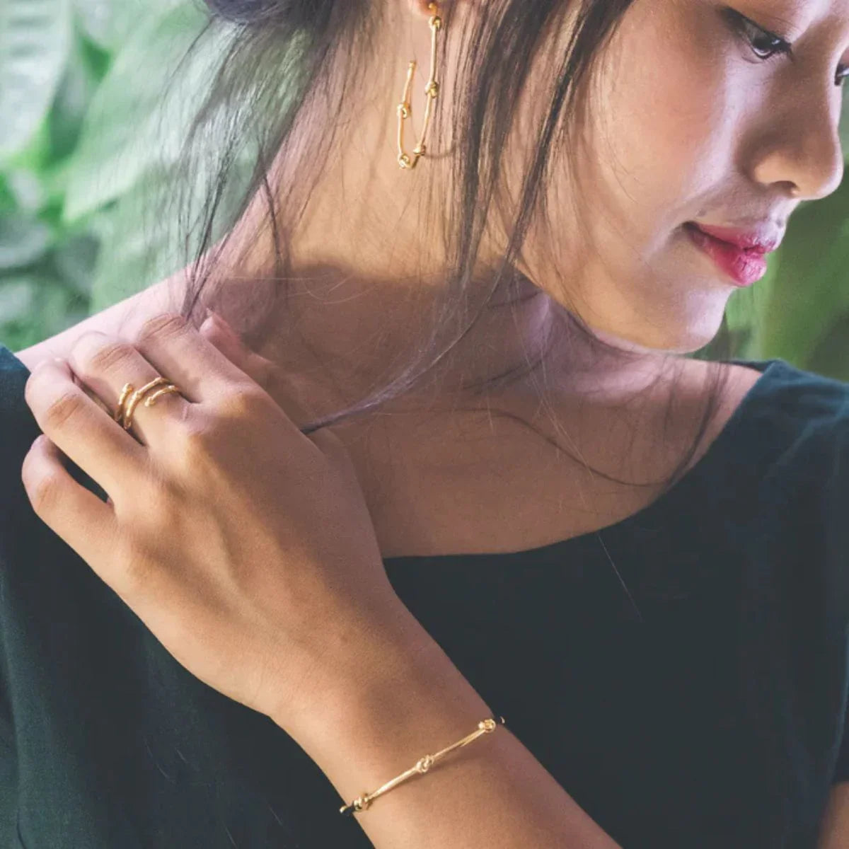 Woman wearing gold jewelry including earrings, ring, and bracelet with a blurred green background