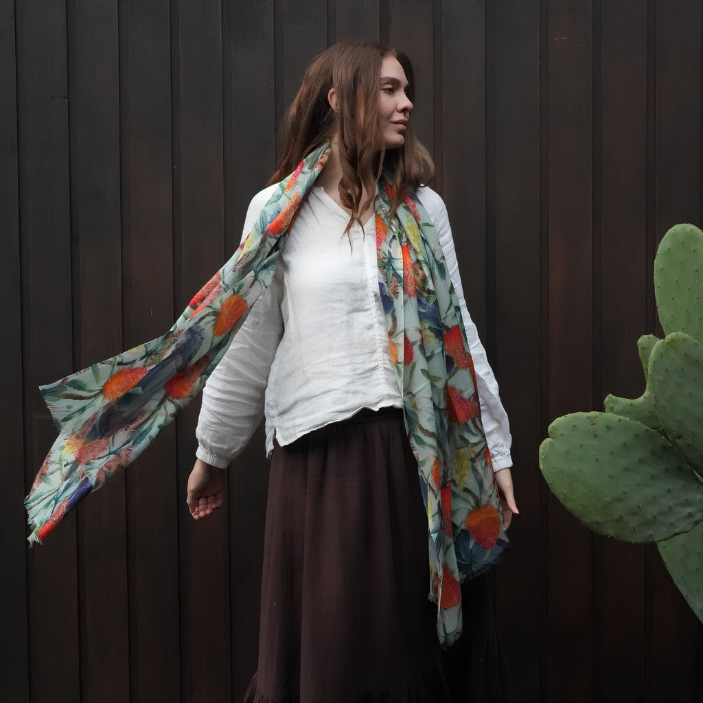 A woman wearing a white long-sleeve top and a dark brown skirt, draped with a lightweight scarf featuring a colorful floral pattern in orange, red, and blue on a light background, standing against a dark wooden plank wall.