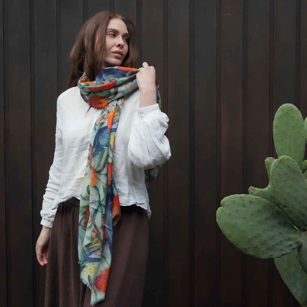 A woman wearing a white blouse, brown skirt, and a colorful patterned scarf stands against a dark brown wooden wall with a green cactus visible on the right.