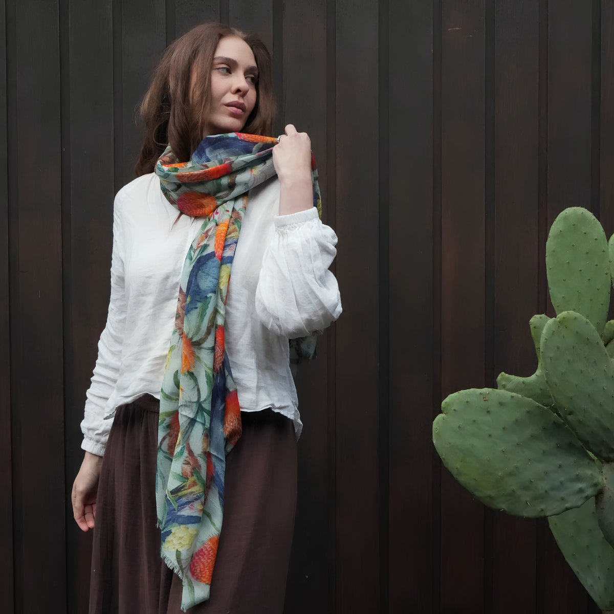 A woman wearing a white blouse, brown skirt, and a colorful patterned scarf stands against a dark brown wooden wall with a green cactus visible on the right.