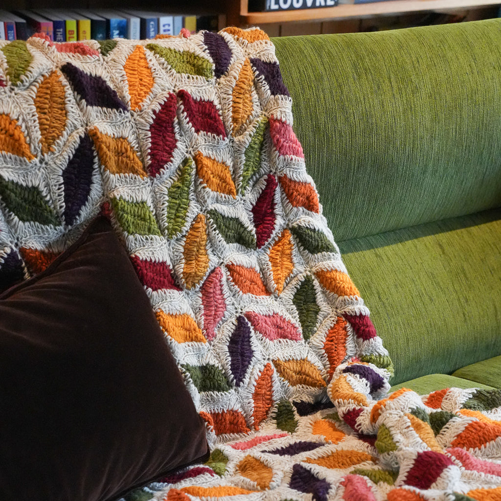 A multicolor textured throw blanket with a leaf-like geometric pattern draped over a green upholstered sofa, with a dark brown pillow in the foreground.