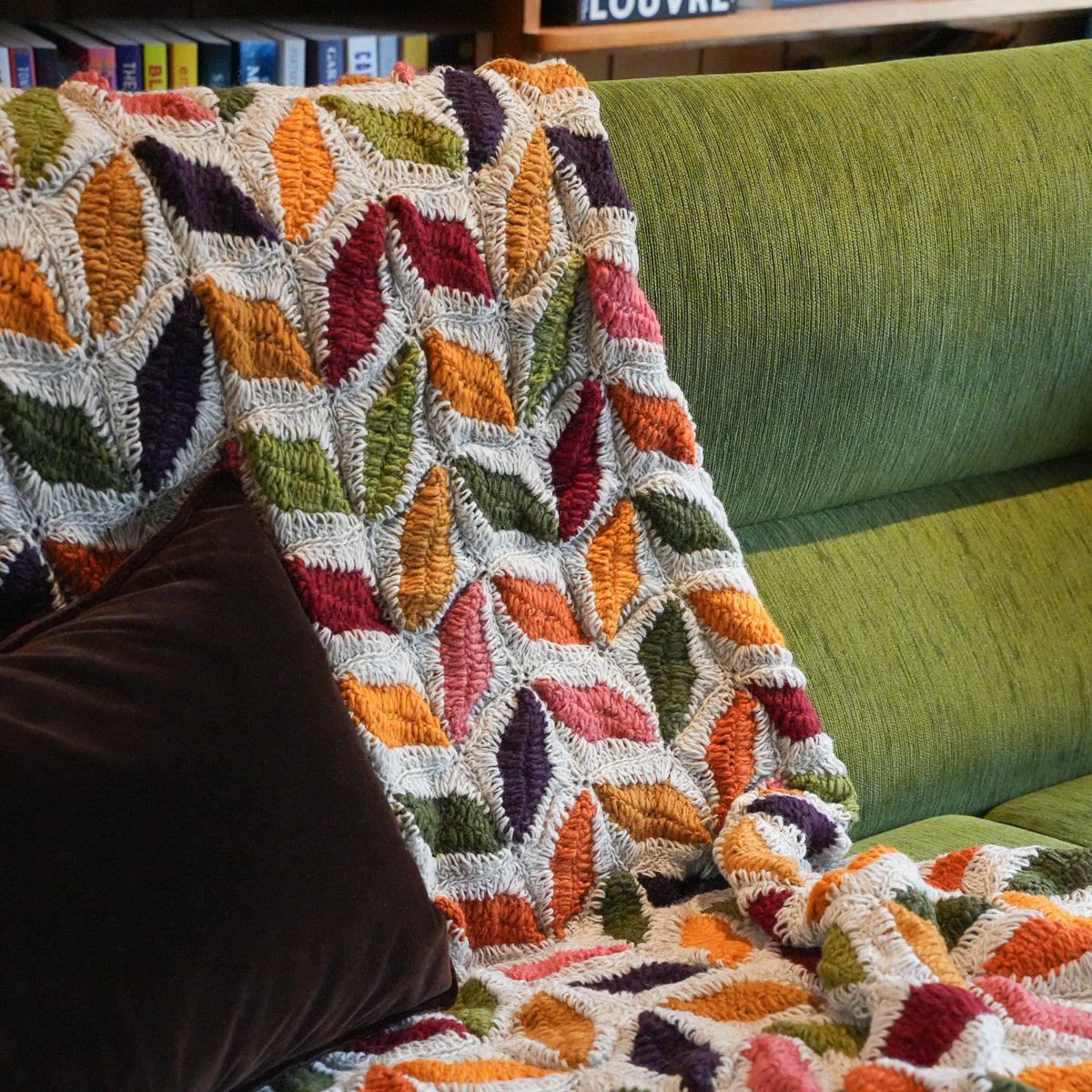 A multicolor textured throw blanket with a leaf-like geometric pattern draped over a green upholstered sofa, with a dark brown pillow in the foreground.