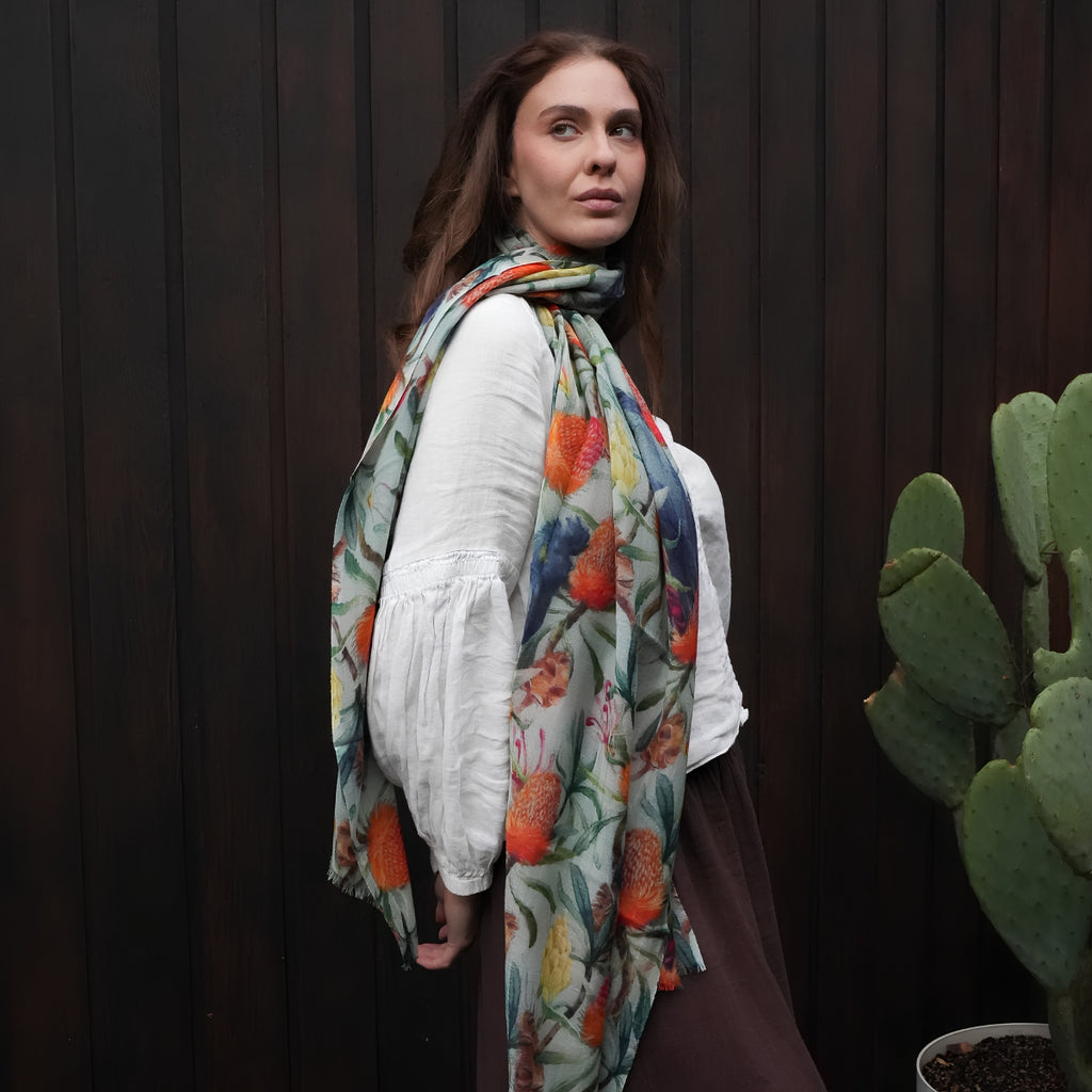 A woman wearing a white long-sleeve blouse and a multicolor floral scarf with orange, red, and blue flowers on a green background, standing in front of a dark wood-paneled wall next to a potted cactus.