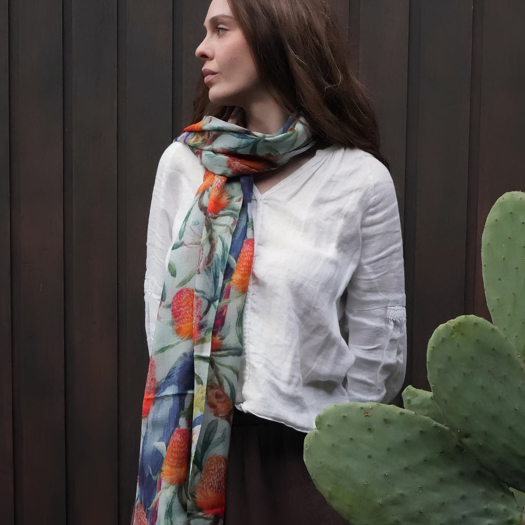 A woman wearing a white linen shirt and a colorful floral scarf with orange, blue, and green tones, standing in front of a dark wood-paneled wall with a cactus in the foreground.