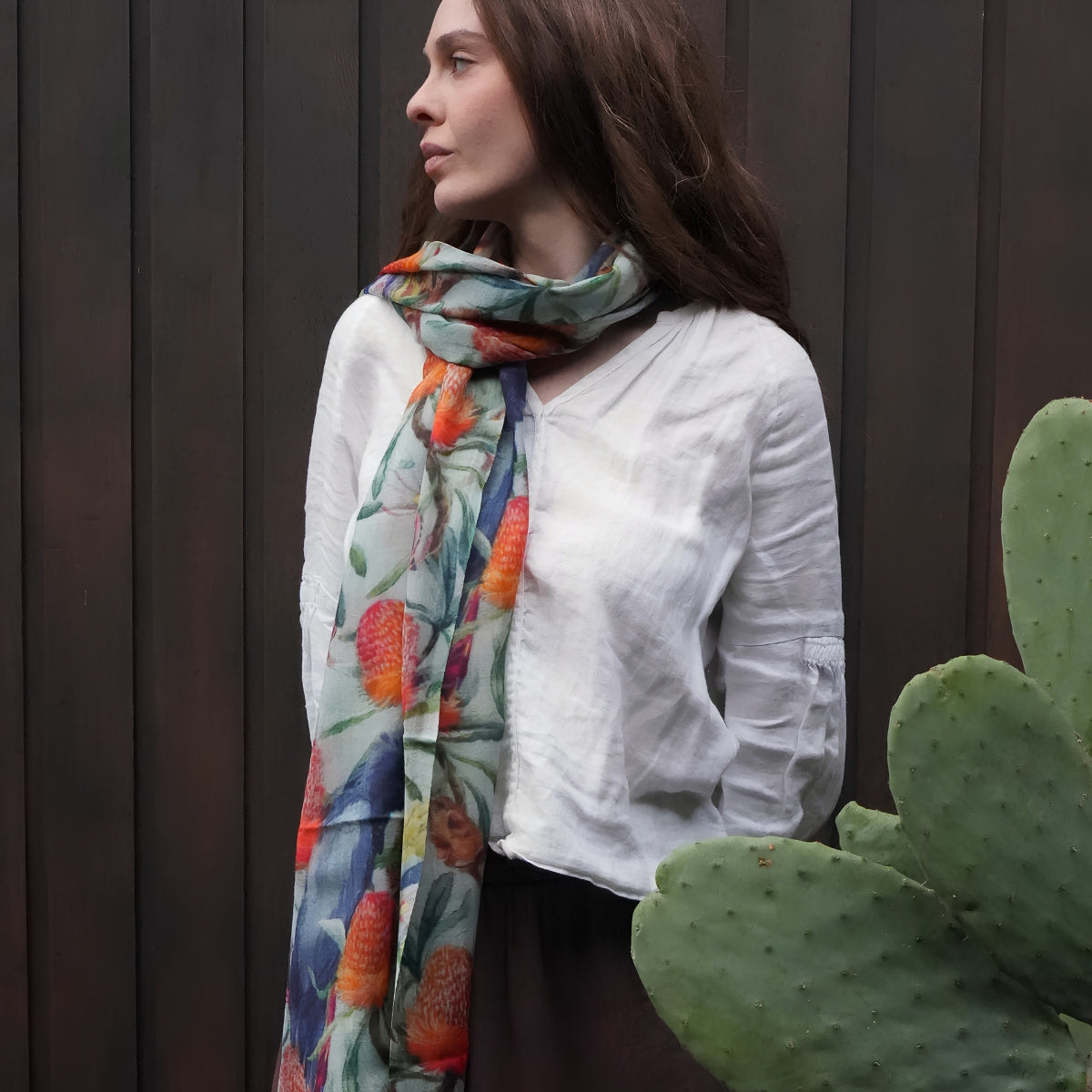 A woman wearing a white linen shirt and a colorful floral scarf with orange, blue, and green tones, standing in front of a dark wood-paneled wall with a cactus in the foreground.