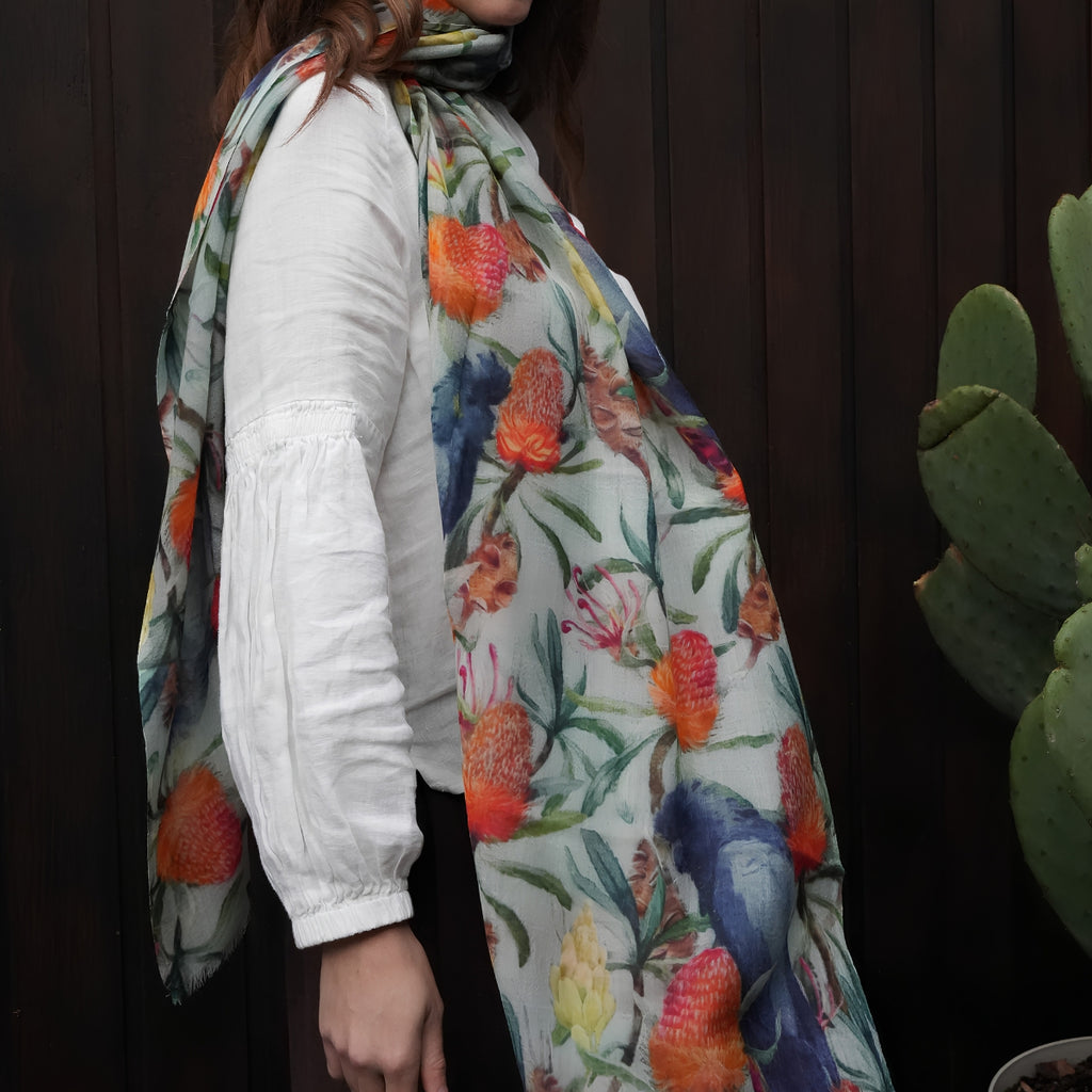 A woman wearing a white long-sleeve top and a lightweight floral scarf draped over her shoulder, shown against a dark wooden background with a cactus visible on the right.