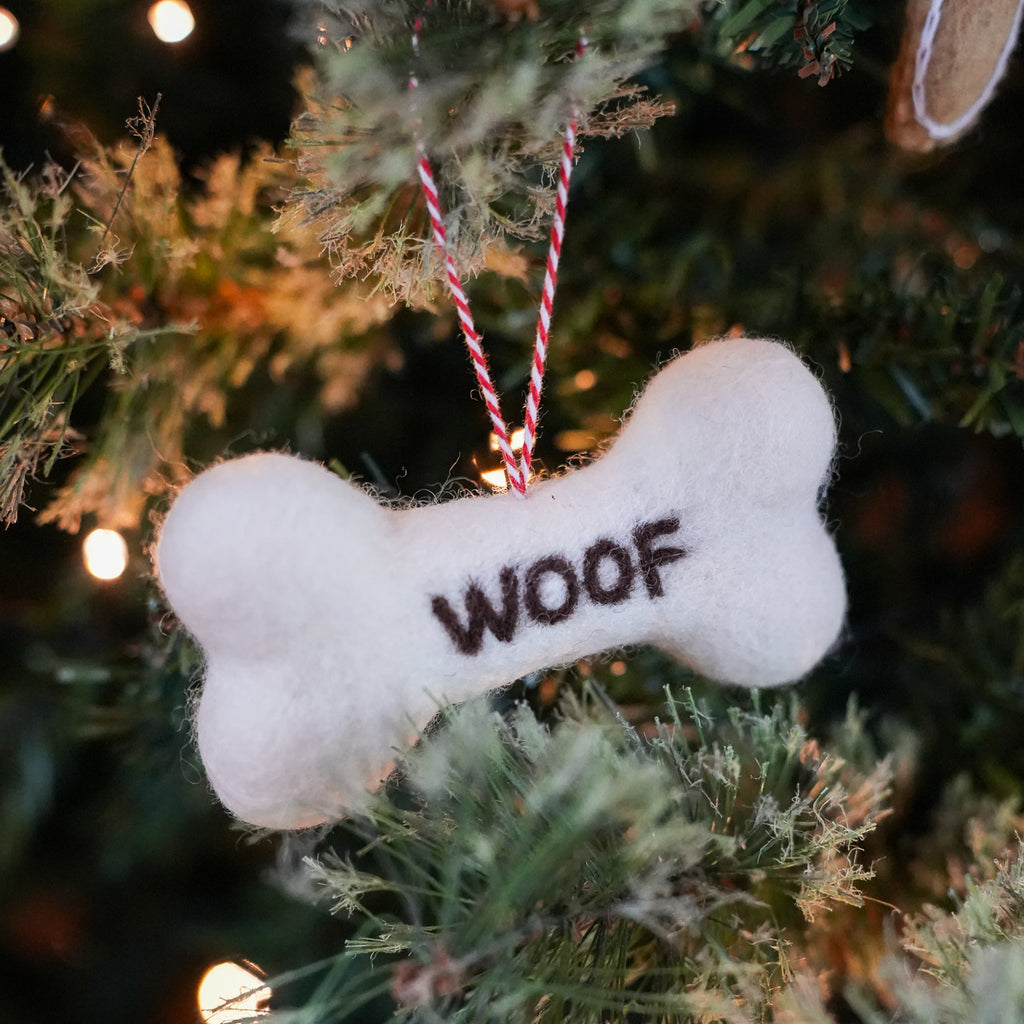 A white felt dog bone ornament with the word 'WOOF' embroidered in dark brown, hanging from a red and white striped string on a decorated Christmas tree.