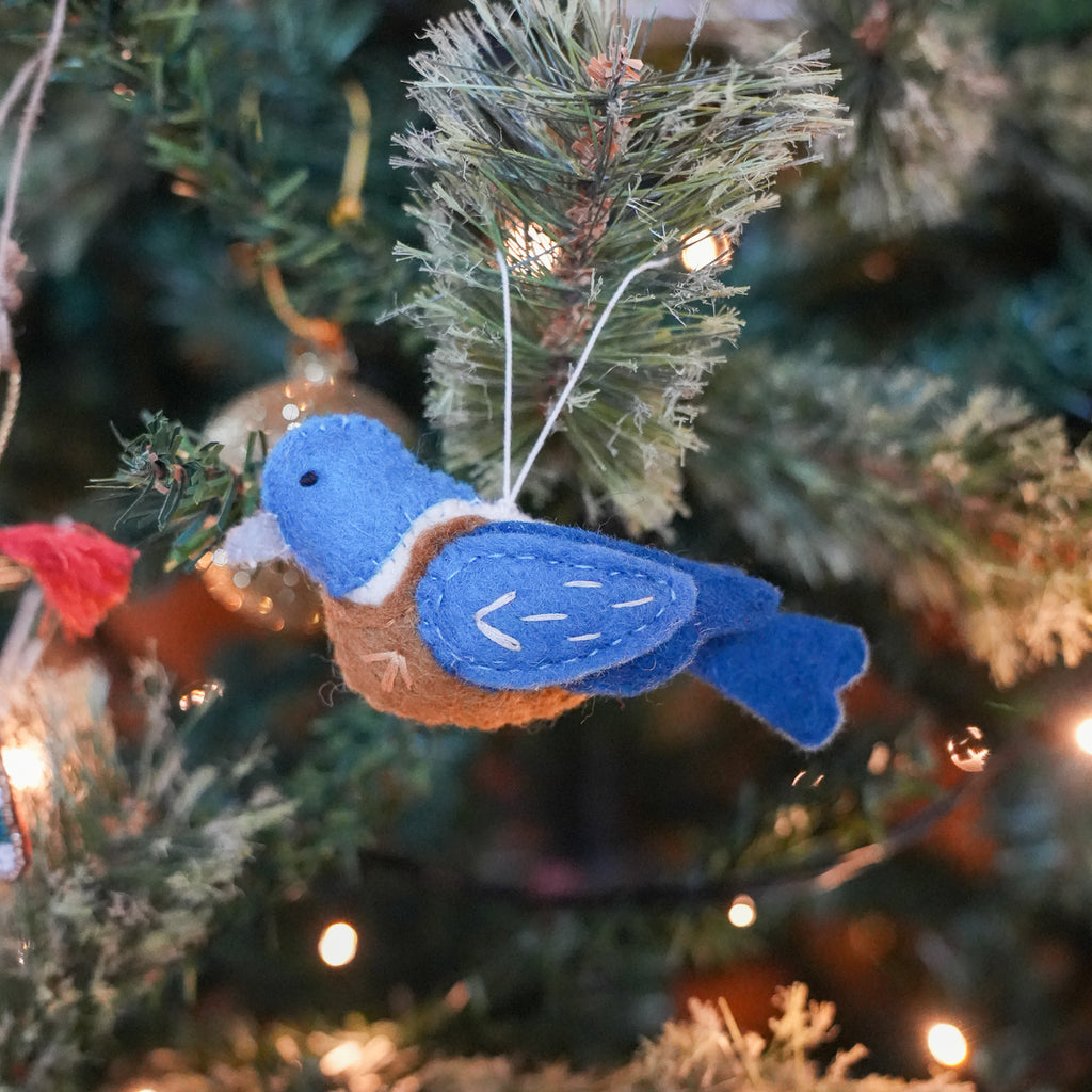 A felt bird Christmas ornament in blue and orange hanging from a decorated Christmas tree with warm string lights.