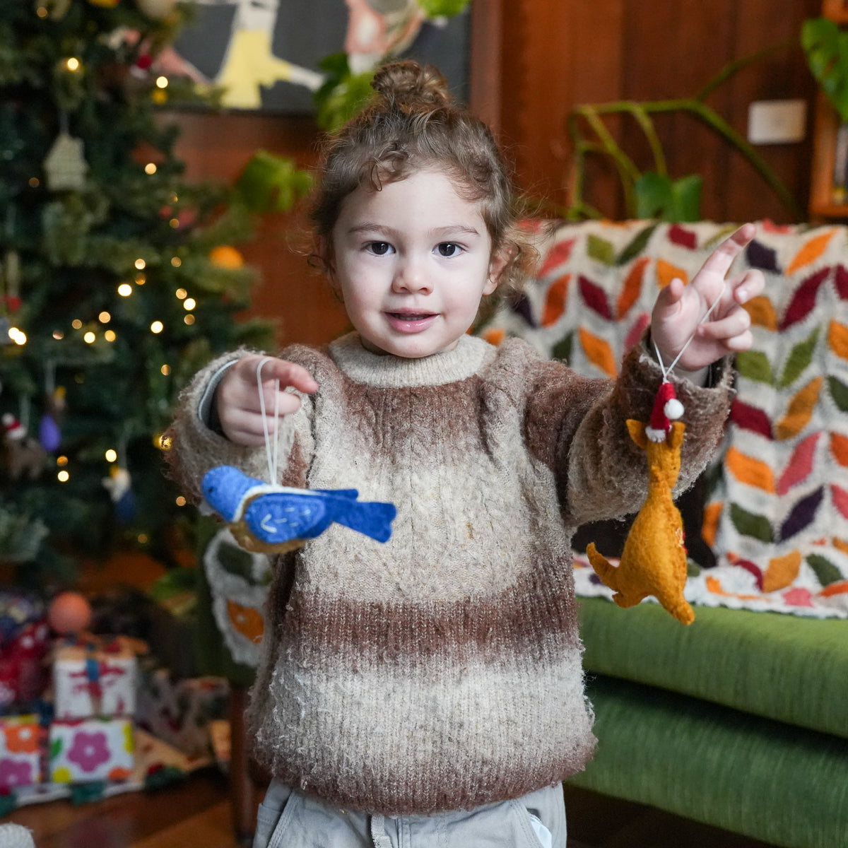 A young child wearing a brown and beige striped fuzzy knit sweater stands indoors holding two felt Christmas ornaments, with a decorated Christmas tree and cozy living room background.