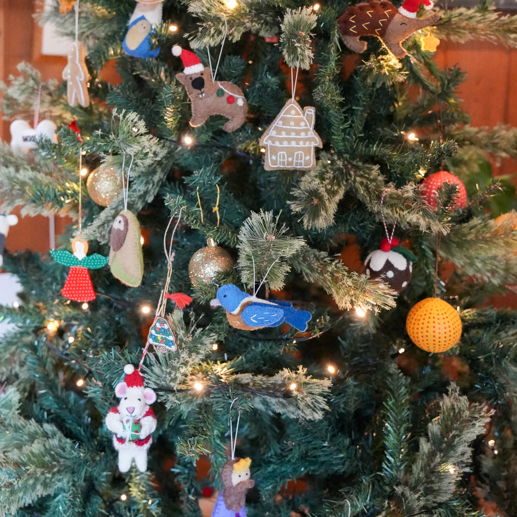 A close-up of a decorated Christmas tree with colorful handmade ornaments, including a blue bird, gingerbread house, teddy bear, and gold and orange baubles, lit by warm white string lights.