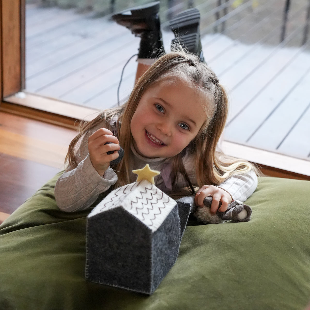 A young girl smiling while lying on an olive green beanbag, interacting with a small black felt house toy with a white patterned roof and a yellow star topper, with a window and deck visible in the background.