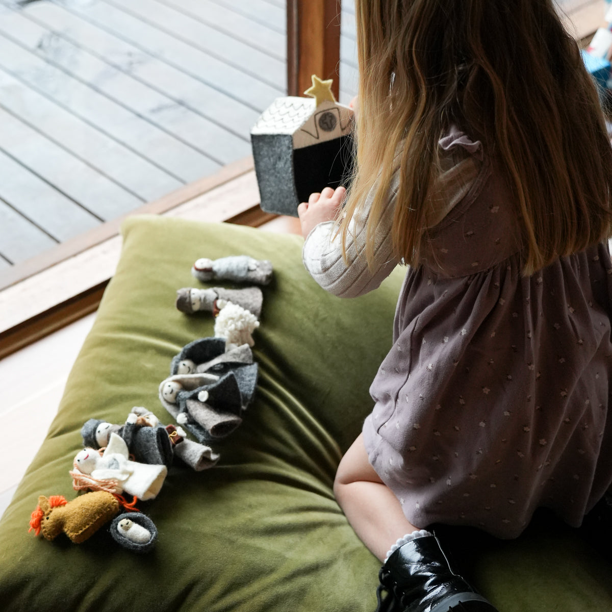 A young child sits on an olive green beanbag, playing with a small wooden house and miniature doll accessories on a wooden deck floor.