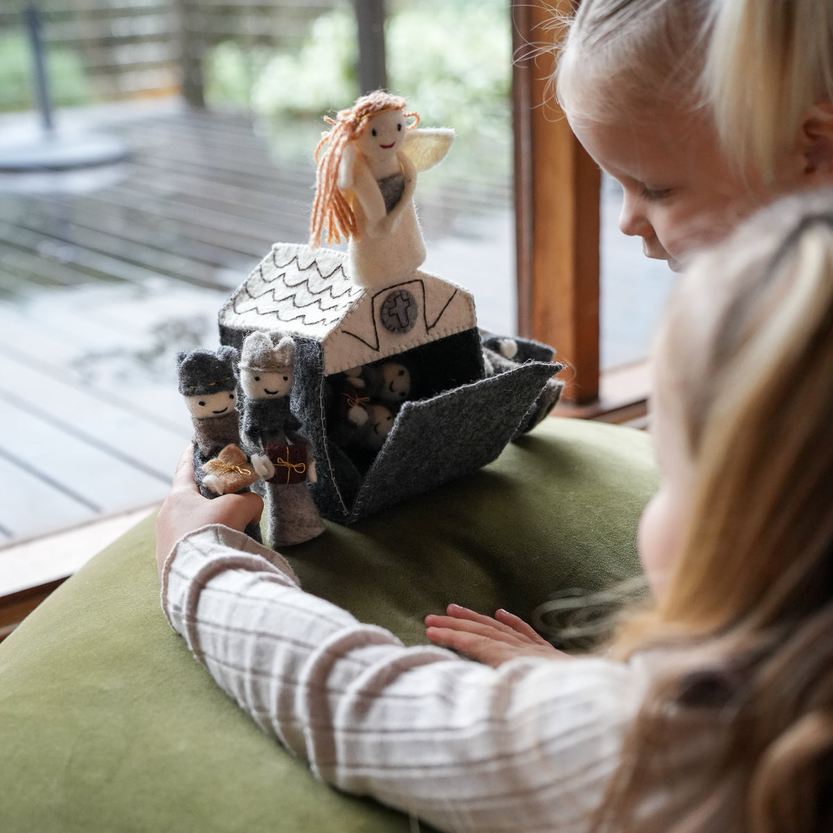 A child holds a small felt house playset with tiny doll figures near a window, with another child leaning in closely. The playset includes a gray felt house, a cream-colored figure with orange hair, and smaller dark and gray characters.