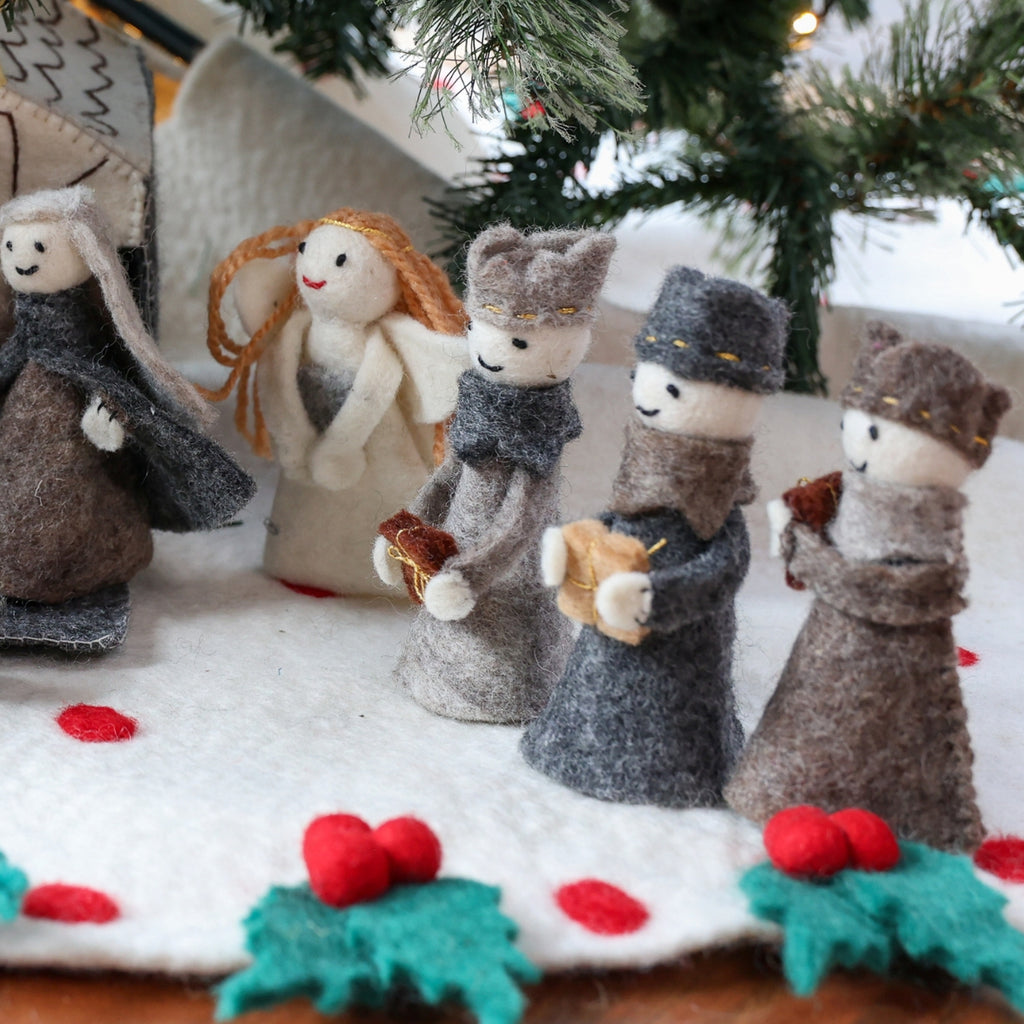 A set of handmade felt Christmas figurines styled on a white fabric surface with holly and a Christmas tree in the background.
