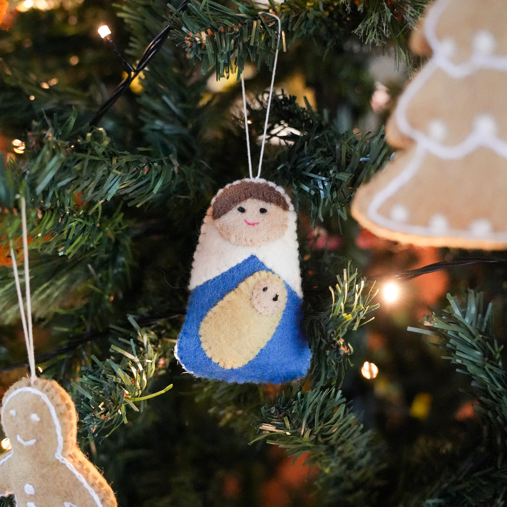 A handmade felt Christmas ornament depicting Mary and baby Jesus hanging on a decorated Christmas tree with warm string lights.