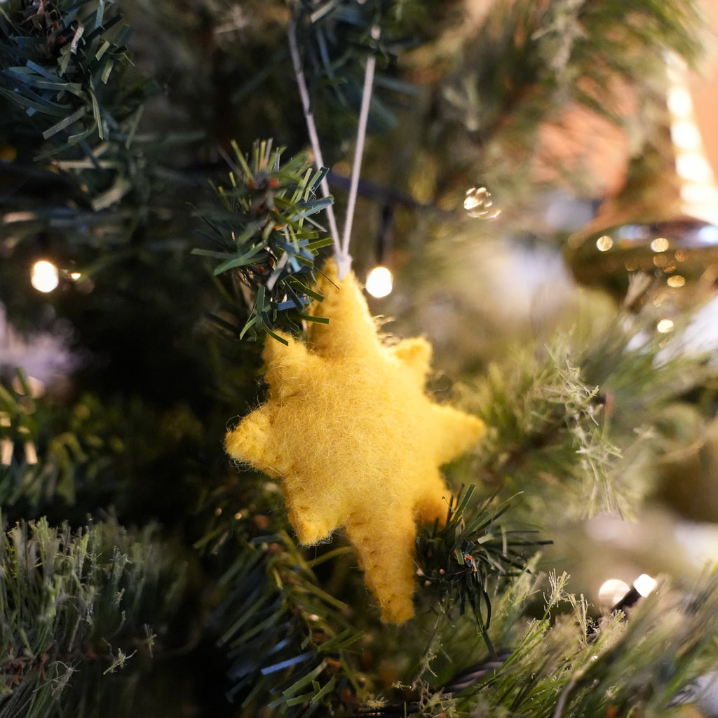 A yellow felt star Christmas ornament hanging from a green artificial Christmas tree with warm string lights in the background.