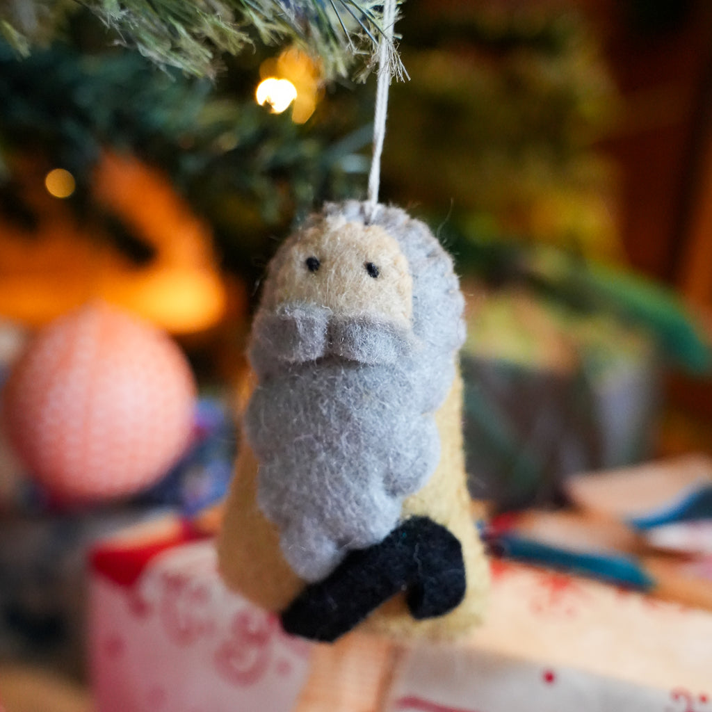 A handmade felt Christmas ornament shaped like a small figure with a gray beard, cream face, yellow body, and black boots, hanging from a Christmas tree branch with a blurred festive background.