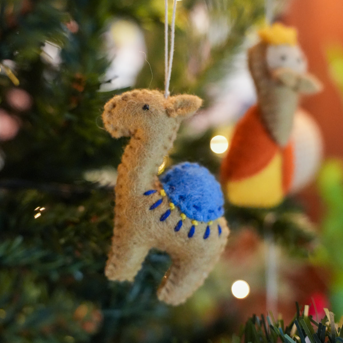 A handmade felt camel Christmas ornament hanging on a green Christmas tree with warm string lights. The ornament is made of tan felt with a bright blue saddle and yellow accents.