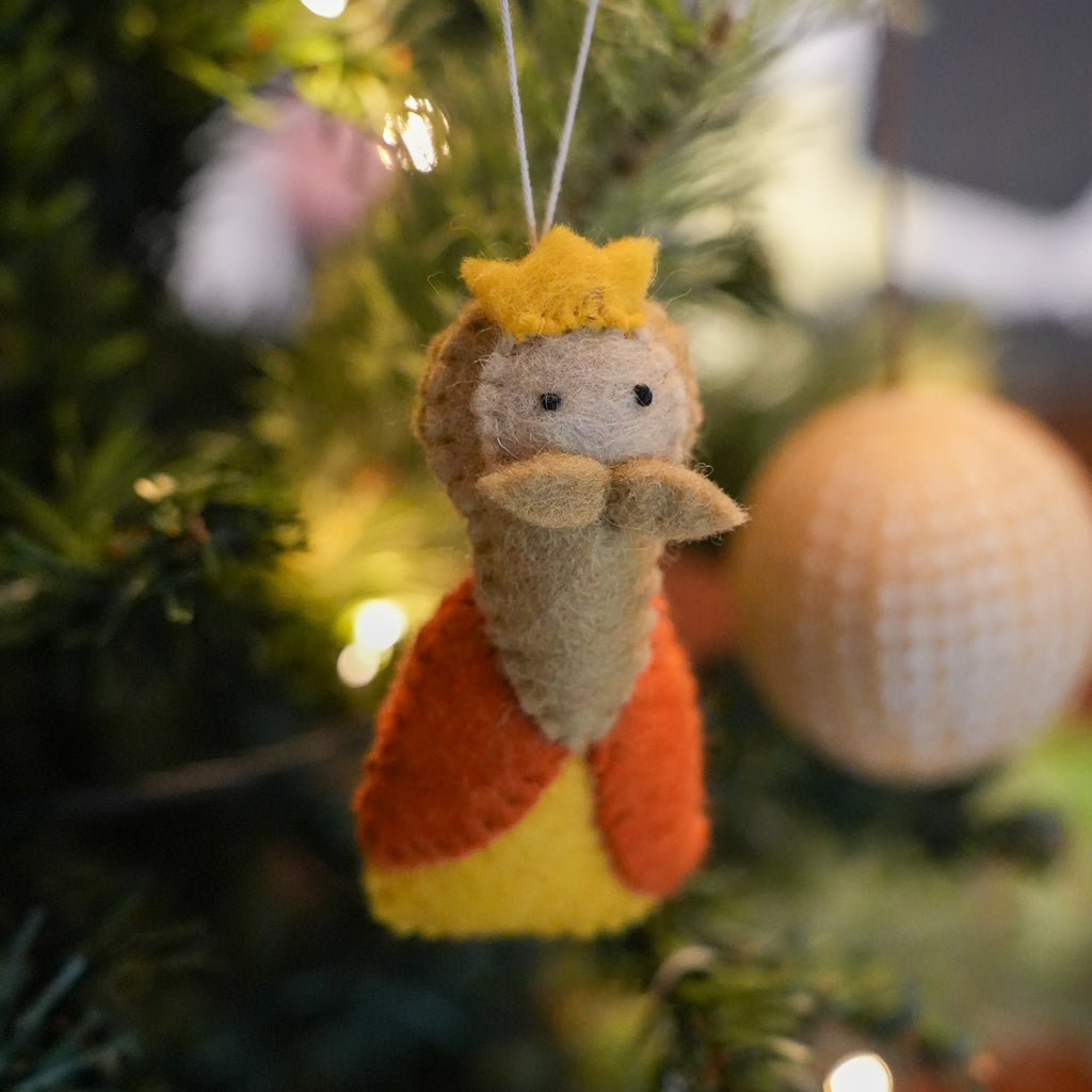 A handmade felt Christmas ornament shaped like a king with a yellow crown, brown hair, and a yellow-and-orange robe, hanging on a blurred Christmas tree with warm lights.