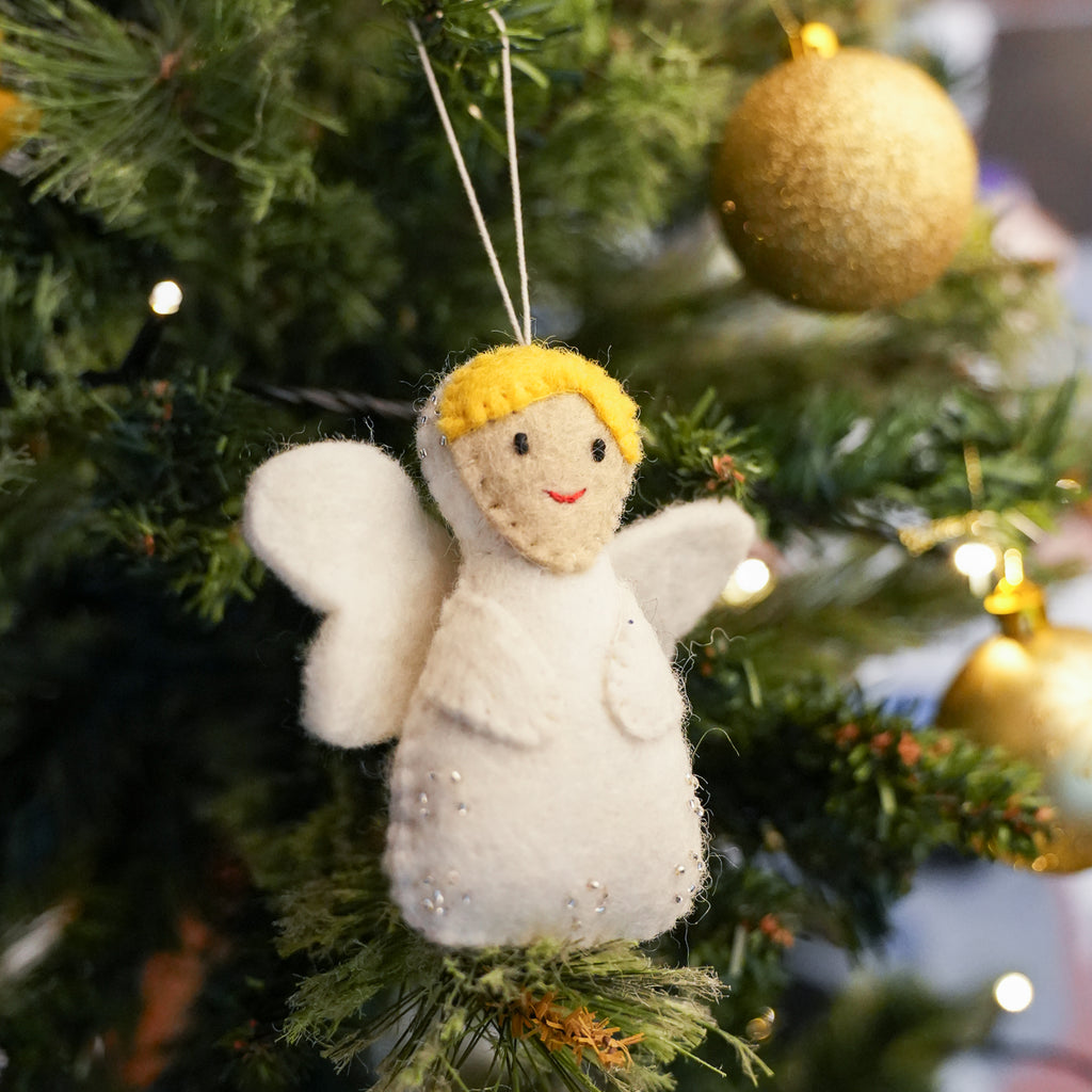 A handmade felt Christmas angel ornament with white wings, a yellow hair, and a red stitched smile, hanging on a green Christmas tree with gold ornaments.