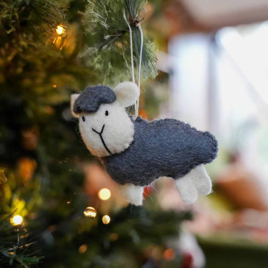 A felt sheep Christmas ornament with a dark gray body and face, and white woolly legs and ears, hanging from a pine branch with warm string lights in the background.