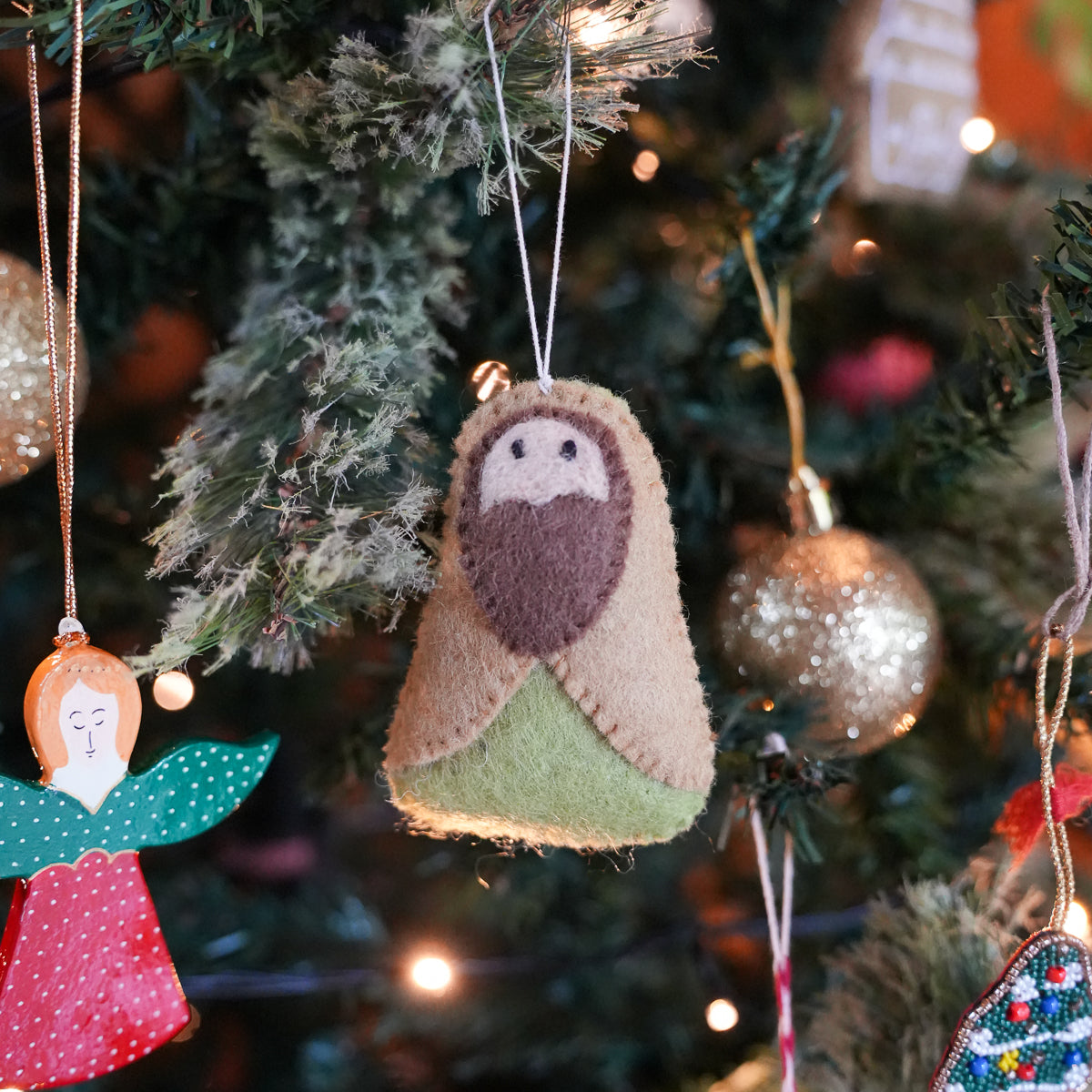 A handmade felt Christmas ornament shaped like baby Jesus hanging on a decorated Christmas tree. The ornament features beige wool for the body, a brown felt beard, and a light green garment, with a white string for hanging.