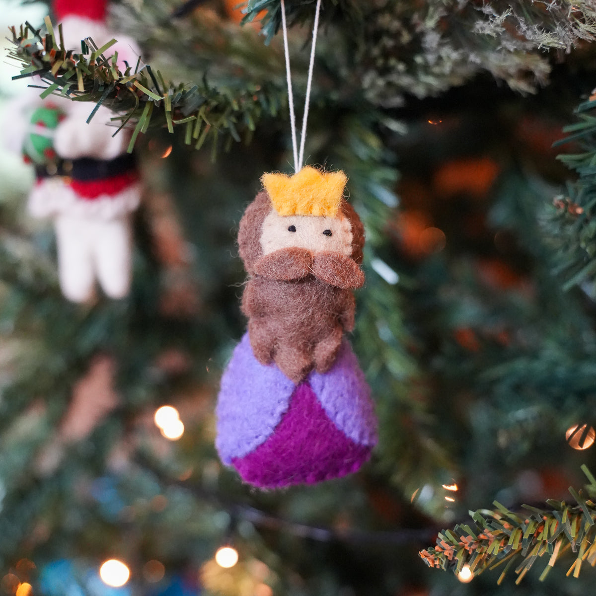 A handcrafted felt Christmas ornament shaped like a king with a yellow crown, brown beard, and purple garments, hanging on a decorated Christmas tree branch with warm lights in the background.